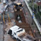 Imagem aérea mostra uma grande vala aberta em uma rua pavimentada, com um homem em pé dentro dela. Nas proximidades, há um caminhão caçamba e uma máquina retroescavadeira