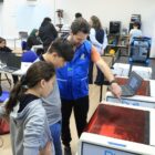 Meninas e meninos adolescentes uniformizados estão em uma ampla sala. Em primeiro plano, um menino e uma menina acompanhados de um professor observam um equipamento