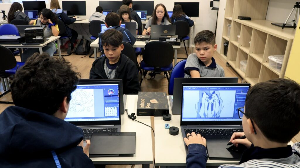 Meninos adolescentes uniformizados estão sentados junto a carteiras e observam atentos as telas de computadores