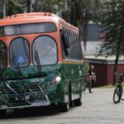 Ônibus nas cores verde e laranja está parado. Ao lado dele, há um homem com uma bicicleta