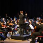 Em um palco, músicos vestidos de preto se apresentam com diferentes instrumentos. Ao centro, uma mulher está virada de costas para a câmera orientando o grupo
