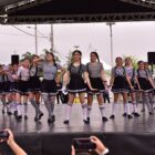 Bailarinas adolescentes vestidas com saias pretas e blusas listradas em branco e preto dançam em um palco aberto