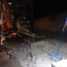 Máquina retroescavadeira está próxima a um buraco no chão. No local, também há um homem uniformizado e com capacete. A imagem é noturna e uma lâmpada ilumina o buraco