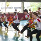 Meninos e meninas adolescentes usando calças pretas e camisetas de uniforme seguram adereços enquanto dançam em uma quadra esportiva