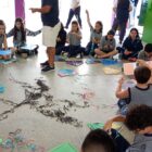 Crianças e adolescentes uniformizados estão sentados em um círculo no chão, produzindo pipas