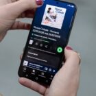Mãos femininas seguram um celular cuja tela mostra a página do spotify do podcast nossa cidade