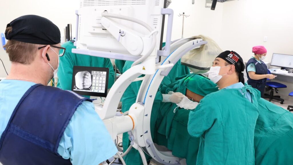 Imagem mostra um paciente deitado de forma lateral, com uma cicatriz recente nas costas. Há dois médicos próximos, que observam o exame de imagem, além de um técnico que opera o equipamento