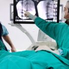 Médicos vestidos com jalecos verdes observam a imagem de raio-x em um equipamento. Há um paciente com o corpo coberto na mesa