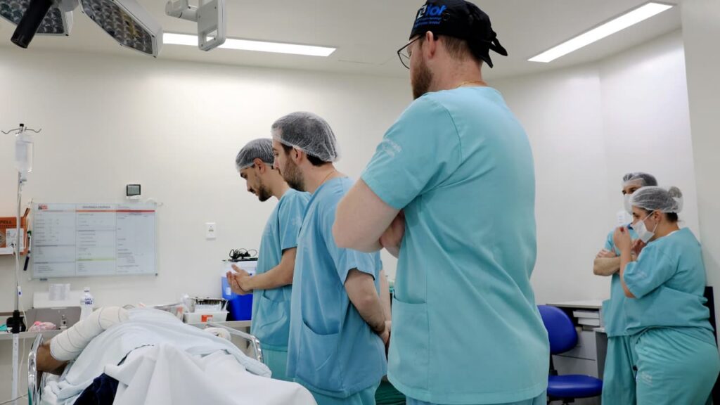 Três homens e duas mulheres vestidos com roupas hospitalares verdes estão em uma sala cirúrgica. Os três homens estão próximos a um paciente que está deitado em uma maca