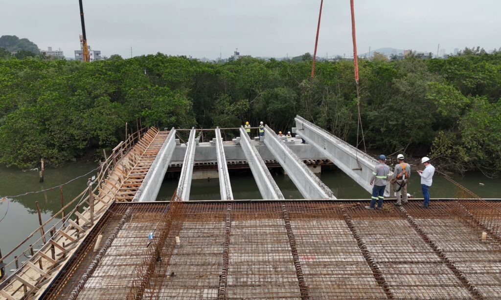Uma viga está sendo içada e posicionada ao lado de outras cinco vigas sobre um rio, na construção de uma ponte