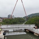 Uma viga está sendo içada e posicionada sobre os apoios de uma ponte em construção