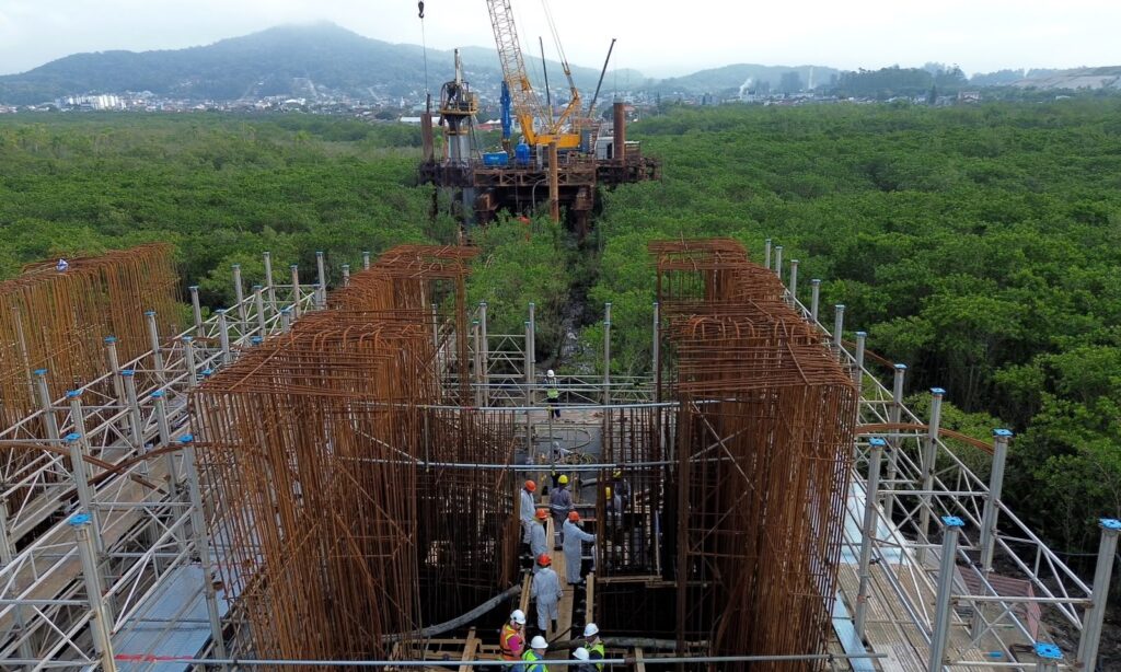 Imagem aérea mostra grande construção, com armação em ferro, guindaste e outros equipamentos, em uma região de rio e vegetação
