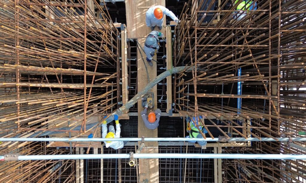 Imagem aérea mostra cinco trabalhadores uniformizados com capacete trabalhando em área com grandes armações de ferro. No local, uma mangueira direciona o concreto para a área da laje