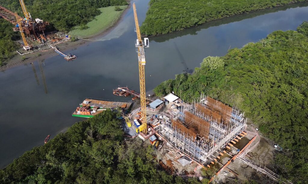 Imagem aérea mostra grande construção, com armação em ferro, guindaste e outros equipamentos, em uma região de rio e vegetação