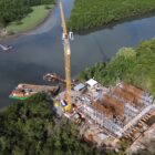 Imagem aérea mostra grande construção, com armação em ferro, guindaste e outros equipamentos, em uma região de rio e vegetação