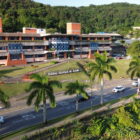 Grande prédio da Prefeitura de Joinville, com tijolos a mostra e detalhes em azul. À frente do prédio, há um amplo gramado e uma rua pavimentada com carros passando, além de palmeiras às margens
