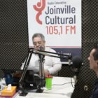 Em um estúdio de rádio, um homem entrevista outro e banner mostra o termo Rádio Joinville Cultural