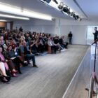 Mulher loira de sobretudo rosa está no palco, em pé, perto de um púlpito. Na foto, é possível ver uma sala ampla, com centenas de pessoas sentadas em cadeiras.