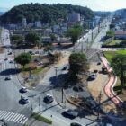 Imagem aérea mostra entroncamento de ruas asfaltadas, com carros passando. Ao fundo há prédios e uma área de vegetação