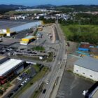 Imagem aérea mostra rua pavimentada com grandes galpões às margens, além de carros e área de vegetação
