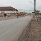 Imagem mostra rua pavimentada com comércio e residências. Ao fundo, há placas indicando obras e um ponto de ônibus
