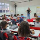 Mulher loira está à frente de sala de aula, com as mãos na cintura. À frente dela, há várias crianças sentadas em cadeiras próximas às carteiras