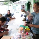 Seis homens de várias idades estão reunidos em uma grande mesa. Em pé, um deles segura um objeto artesanal, enquanto os demais observam