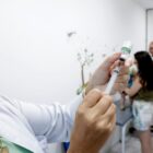 Mulher de jaleco branco segura uma seringa e agulha. Ao fundo, há um homem, uma mulher e um bebê
