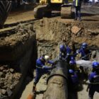 Trabalhadores uniformizados e com capacete trabalham no reparo de uma grande adutora. O tubo de 600 mm, assim como os trabalhadores, estão em uma grande vala aberta em um terreno a céu aberto