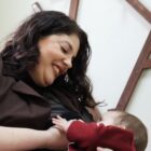 Mulher está sentada em uma poltrona amamentando uma bebê. Ela sorri enquanto olha para a bebê