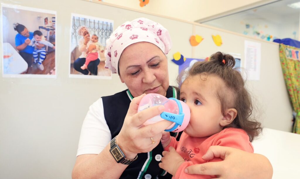 Mulher com touca na cabeça está dando leite em um copo infantil para uma menina de cerca de um ano