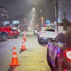 Imagem mostra rua pavimentada à noite, com cones no centro da via, uma viatura da guarda municipal e outros carros