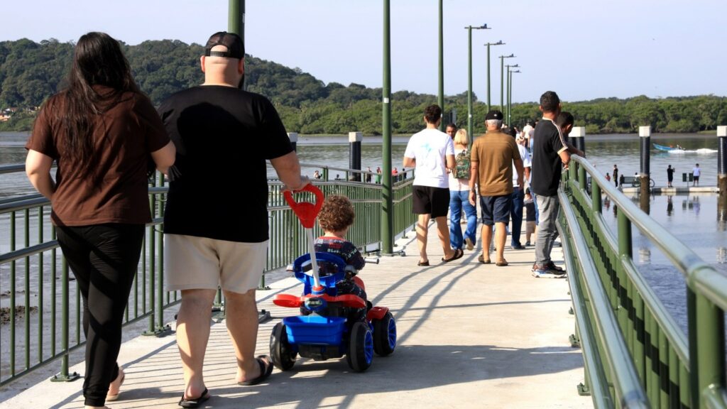 Em primeiro plano, um casal e uma criança em uma motoquinha andam pelo trapiche. Ao fundo, mais pessoas de várias idades fazem o mesmo percurso