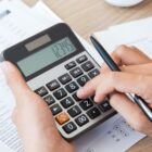 Imagem mostra duas mãos, uma delas segurando uma calculadora e a outra teclando números na calculadora enquanto segura um lápis