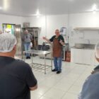 Em uma cozinha com as paredes pintadas de branco e equipamentos em inox, um homem está de jaleco e toca no centro. Ao redor dele, há outros homens ouvindo o que ele fala