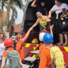 Homem vestido de laranja e usando capacete dá a mão a uma mulher que está descendo da carroceria de um caminhão. Há outras pessoas no chão e no veículo