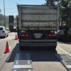 Caminhão caçamba está parado após passar por balança móvel de passagem. Ao lado dele, há um carro do Detrans, além de um guarda municipal e uma agente de trânsito