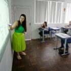 Mulher de óculos com saia e blusa verdes está à frente de sala de aula, escrevendo no quadro. À frente dela, há quatro mulheres sentadas em carteiras prestando atenção
