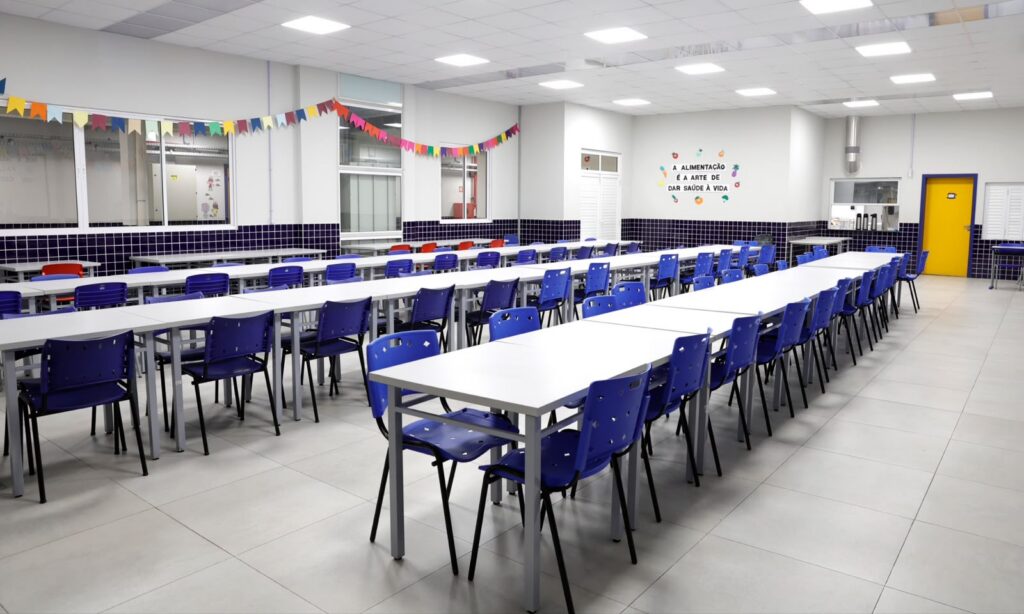 Espaço mostra um amplo refeitório com duas mesas compridas e várias cadeiras em ambos os lados das mesas