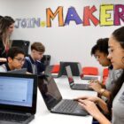 Em uma sala, quatro alunos manuseiam notebooks em uma mesa. Em pé, uma professora observa