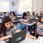 Alunos uniformizados estão em uma sala de aula. Cada um em uma cadeira, eles manipulam notebooks em cima das carteiras. Ao fundo da sala, uma mulher professora fala
