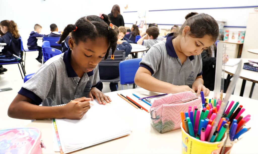 Duas meninas, uma negra e uma branca, estão sentadas em cadeiras escrevendo sobre carteiras. Nas carteiras, há lápis, canetas, um penal entre outros materiais. Ao fundo, há outros alunos na sala