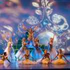Doze meninas de cerca de dez anos estão com figurino que remete a princesas em um palco. Elas fazem uma das poses da coreografia de balé. Ao fundo, há um painel que mostra uma coroa