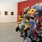 Imagem mostra buquê com flores de diferentes cores em primeiro plano. Ao fundo, há quatros expostos em duas paredes