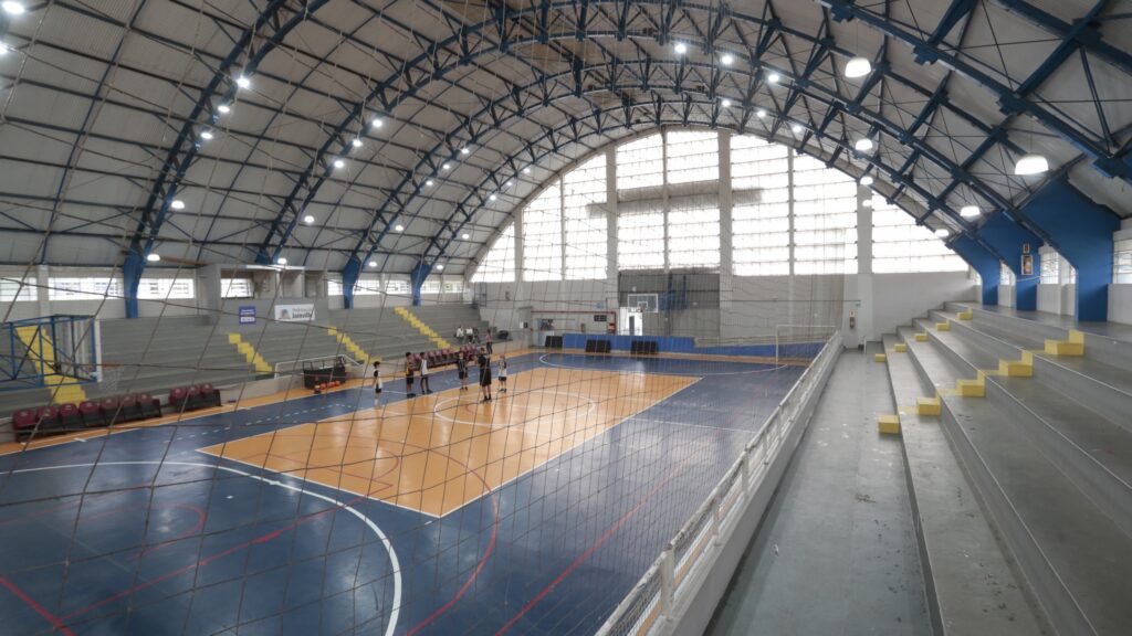 Imagem interna do Ginásio Abel Schulz mostra a quadra de esportes, parte das arquibancadas e cobertura em forma de arco