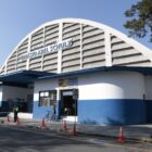 Imagem mostra o Ginásio Abel Schulz, uma grande estrutura de concreto com telhado em forma de arco e pintado nas cores azul e branco