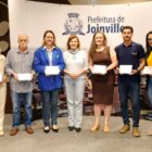 Sete pessoas, cinco pessoas e dois homens, estão em pé, em frente a um painel da Prefeitura de Joinville. Todos sorriem, e com exceção da prefeita ao centro, seguram placas de homenagem
