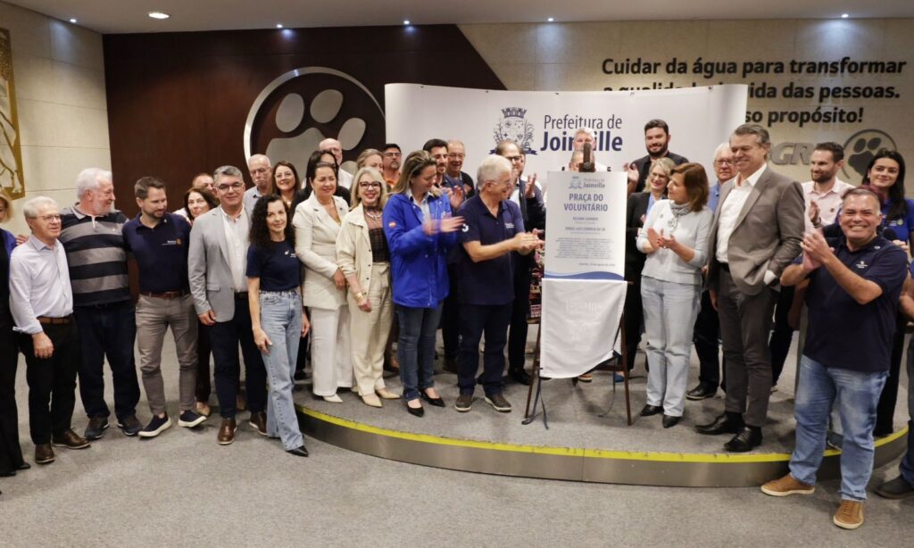 Em uma sala, há mulheres e homens reunidos em pé, sorrindo. No centro do palco, há uma placa metalizada