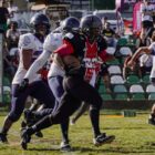 Homens uniformizados e usando capacete jogam futebol americano em um campo de grama com arquibancada ao fundo