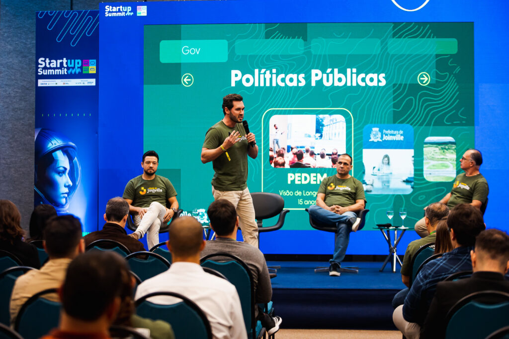 Em um palco, um homem de camiseta verde está em pé, falando no microfone. Outros três homens estão sentados no palco